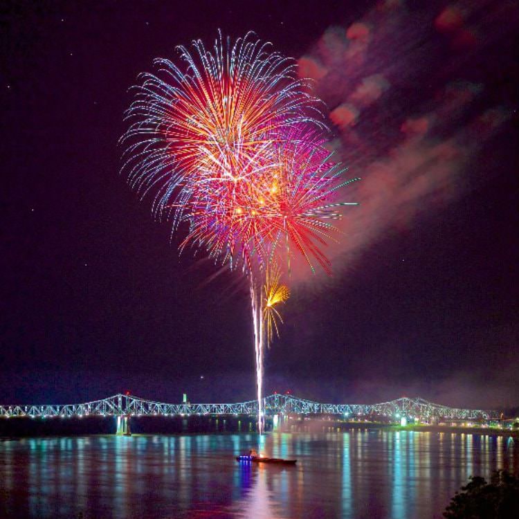 A rainbow colored fireworks display shooting from a small boat over the water with the bridge in the background well-lit at night