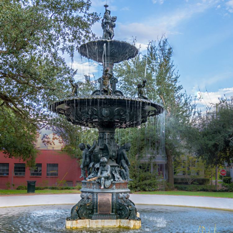 A tall elegant water fountain decorated with cast iron children along the sides spouting water into a large pool with red building behind