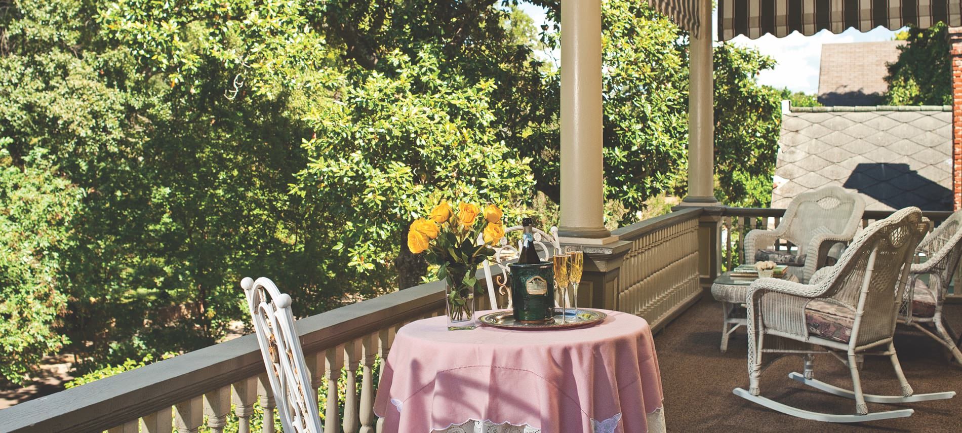 A balcony decorated with cream fencing and pillars, white wicker chairs and a pink tablecloth covering a round table with yellow roses and champagne