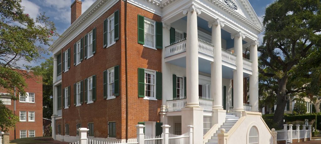 Angled view of a three story b&b made of brick and white siding, green shutters surrounding many windows, and two staircases leading to front porch