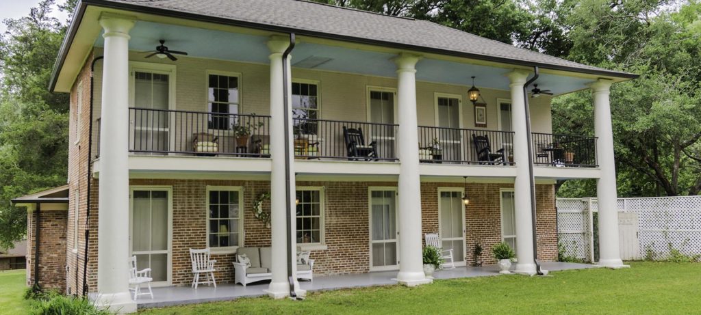 Rear view of the b&b with wide open porch spaces, various chairs and plants, and many windows with a green lawn