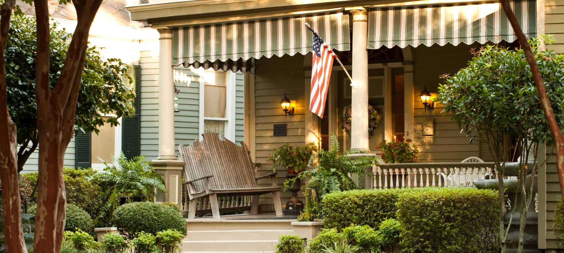 View of front porch covered in ferns and greenery, a rustic bench and furniture, and tan house siding
