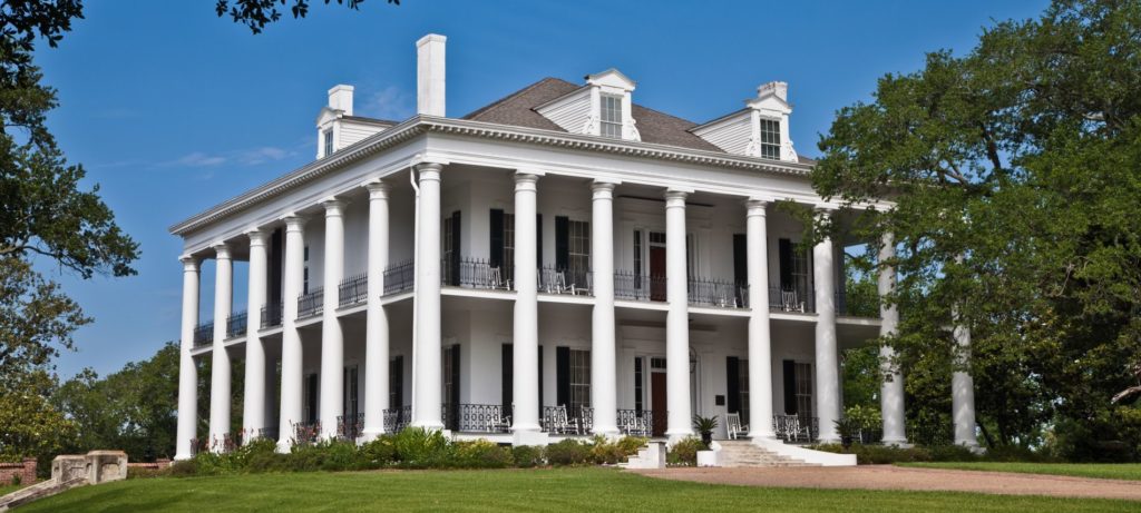 Angled view of two story b&b with white siding, tall white pillas surrounding house, and many windows; grass is green and bright blue sky