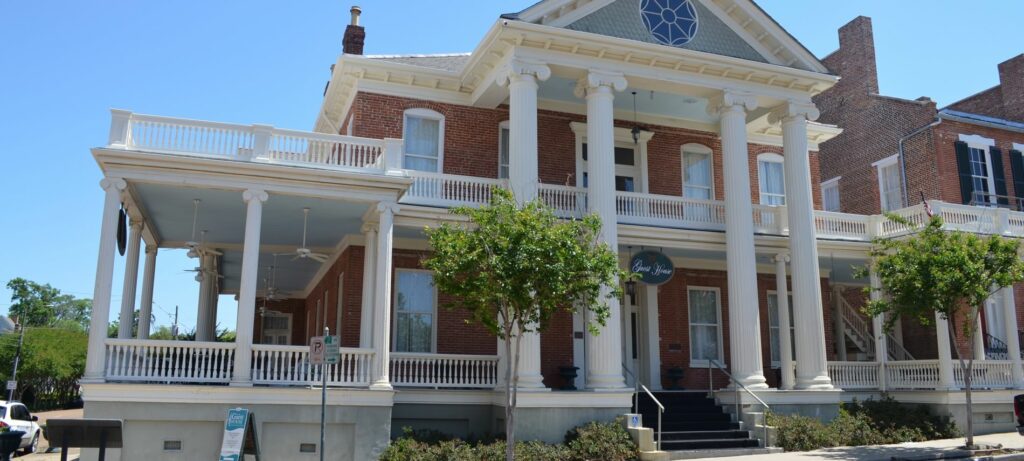 Front view of Guest House that is two stories and covered in red-brick. Has many white pillars, large porch areas, and small trees on front sidewalk