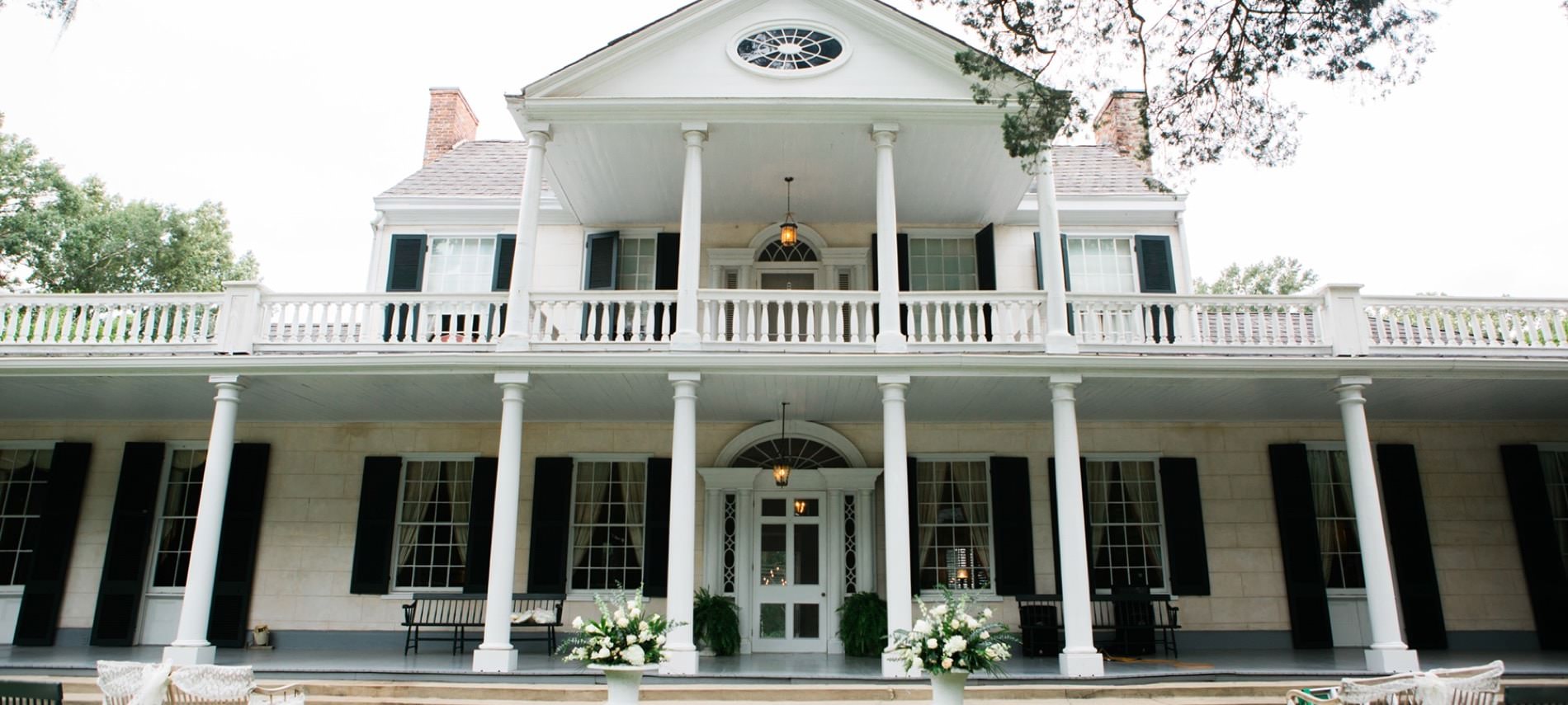 Front view of two story b&b with white stone siding, dark shutters around many windows, white pillars, and intricate white front door
