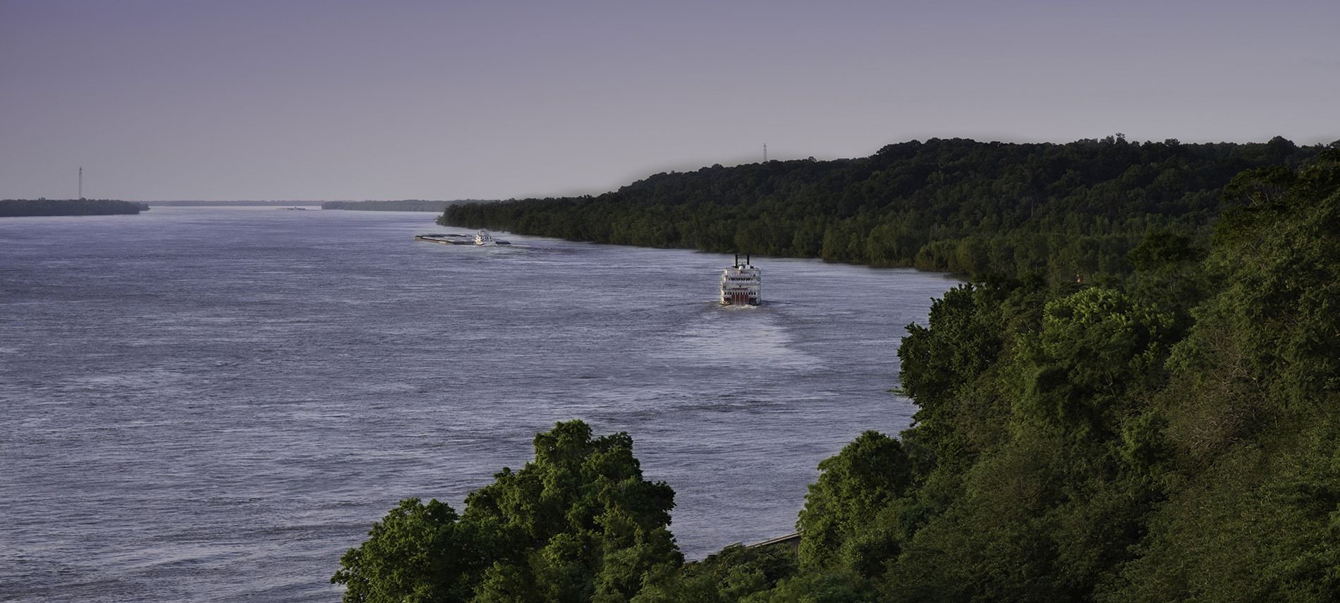 View of trees lining the shore of a lake with a ferry boat riding down the water on a cloudy day