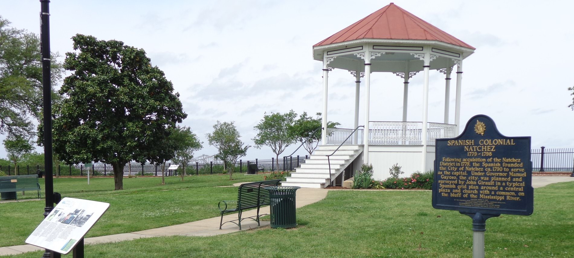 A white gazebo with a red roof overlooking a nice green park with park benches and a pale blue sky