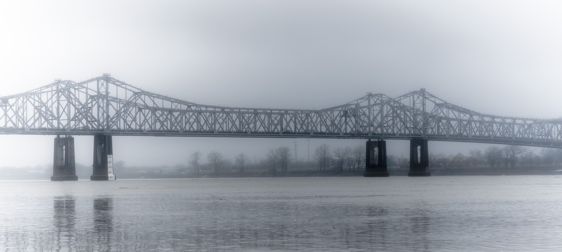 Long driving bridge over the water on a foggy day with bare trees lining the shore in the background