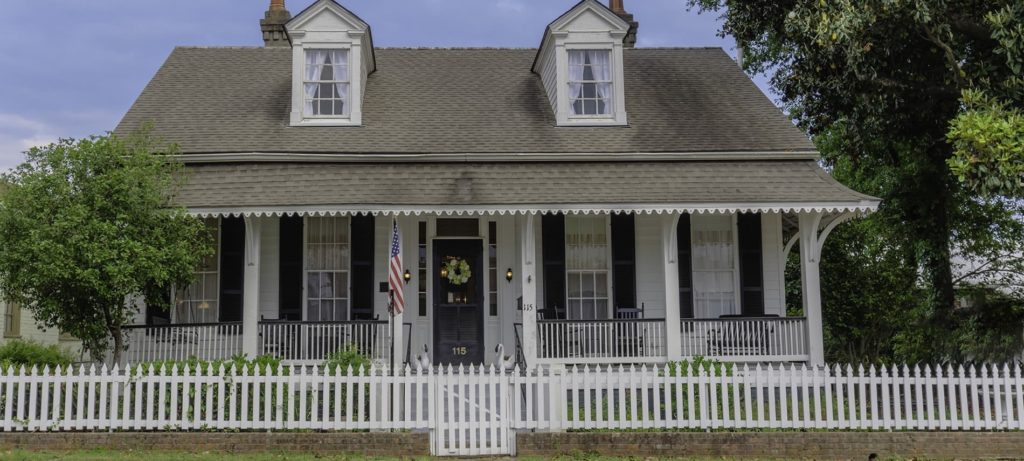 Front view of b&b with short, white fencing, white siding on house with black shuttered windows and front door, slanted roof with two windows, and trees surrounding house