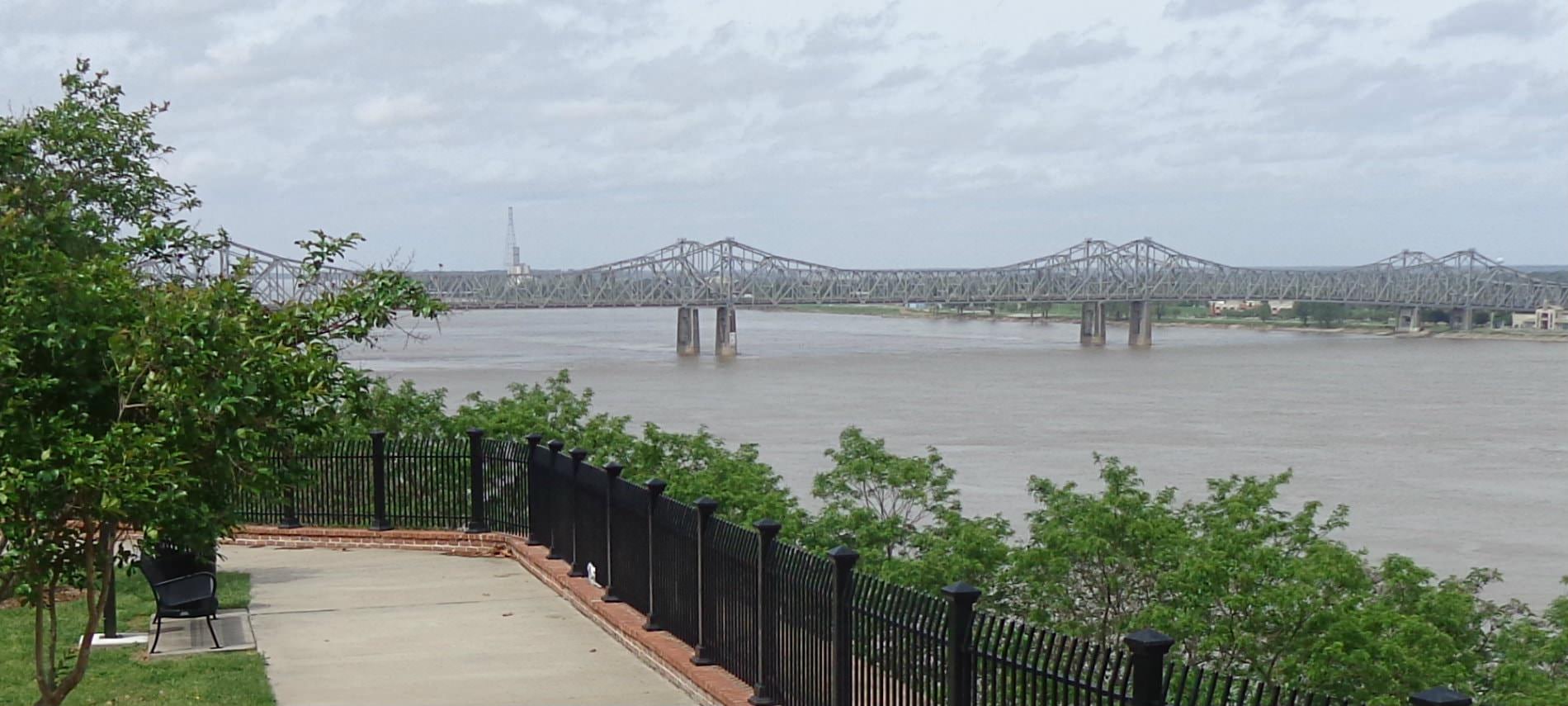 View of a sidwalk in the park overlooking water on a cloudy day and a long bridge in the distance