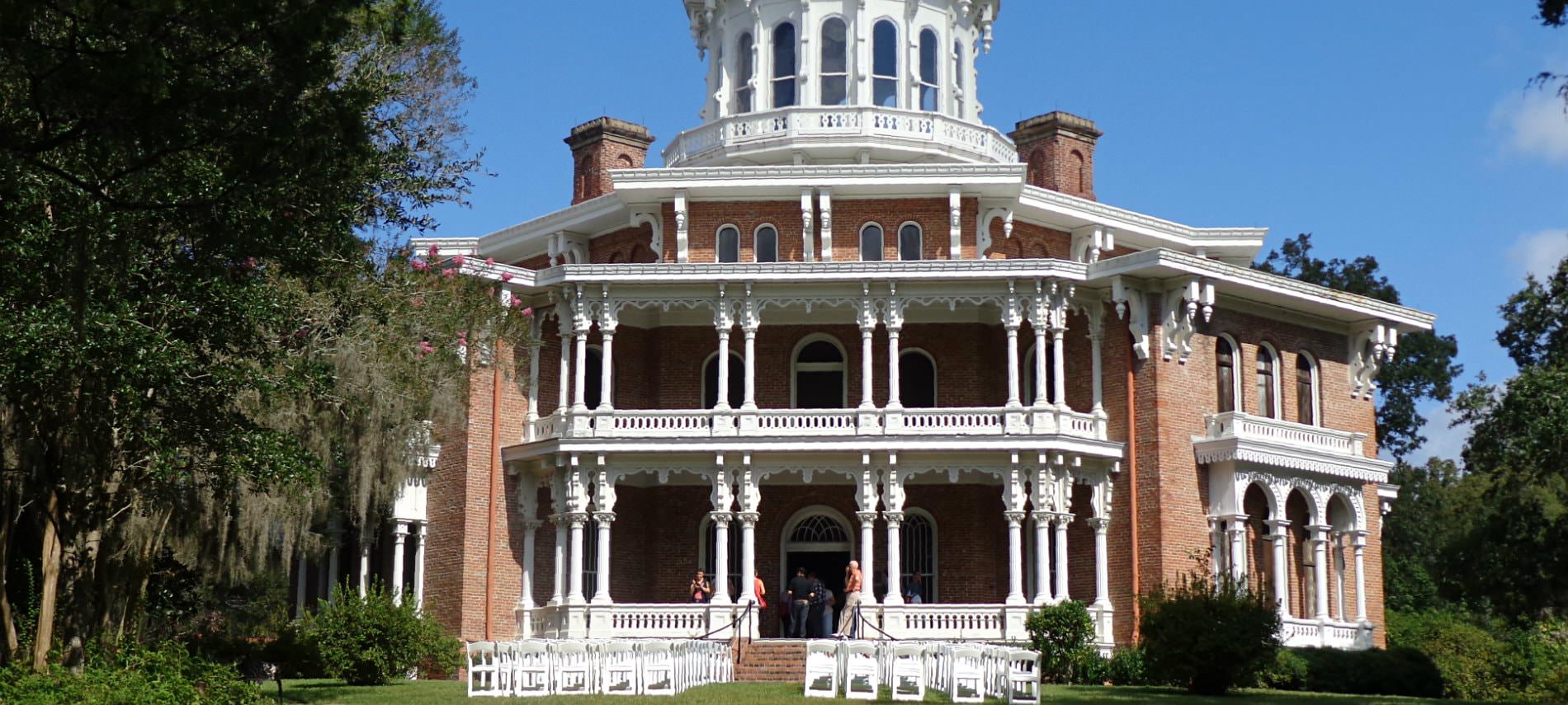 View of a three story brick venue with decorative white pillars and fencing with white chairs in rows in front yard