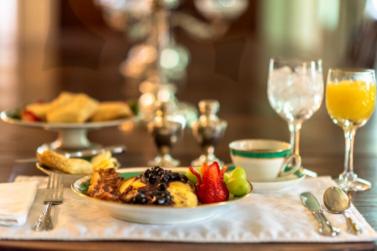 A breakfast table with cloth placemats and napkins, sliverware, a plate full of pancakes covered with a blueberry sauce, bacon, and fresh fruit, along with glasses of orange and water, and a cup of coffee.