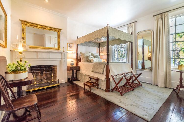 A bedroom with a four poster canopy bed, rich dark wood floors, wood furnishings, a fireplace, a gilded mirror above the fireplace, and tall windows with views of the outdoor trees and flowers.