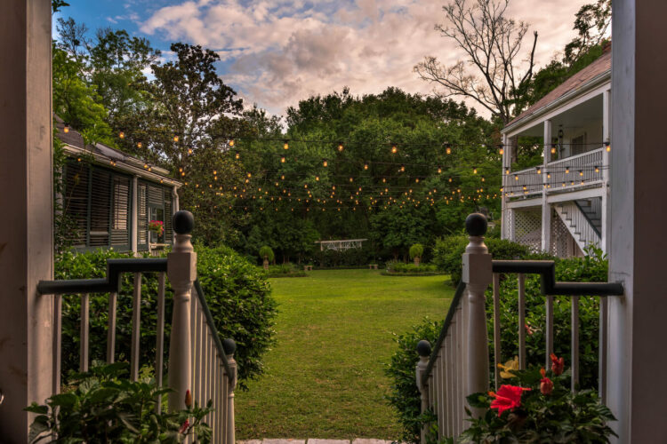 A background with parts of a stately house on either side, a porch going down to a large yard of green grass, a gazebo at the far end of the yard, and twinkle lights strung along the top of the porch.