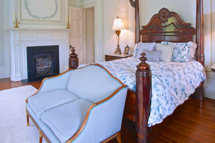 a large bedroom with rich wood floors, an antique wood bed, a blue loveseat, side tables with lamps, a fireplace, and an area rug.