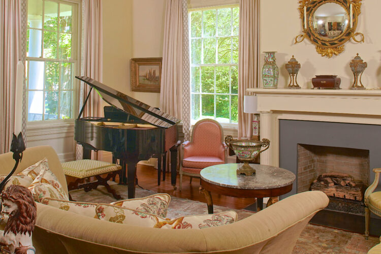 A living room with beige walls, a fireplace with a grey surround and a mantle with ornate vases and decor, a decorative gilded mirror above the fireplace; tall windows with tan curtains, a comfortable couch and some armchairs; and a baby grand piano in the corner.