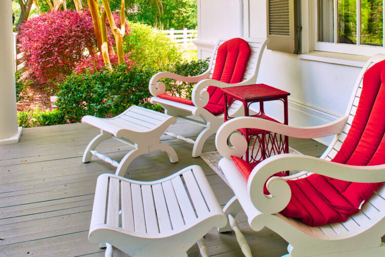 A porch with painted white chairs and red cushions with lush greenery and pink flowered bushes.