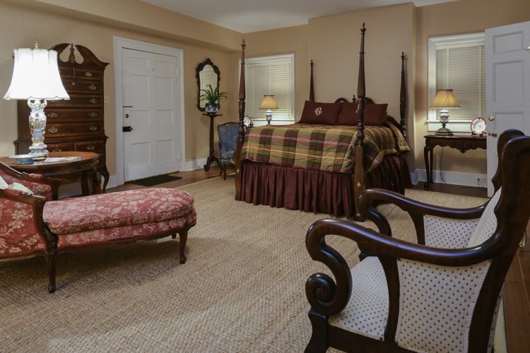 Large bedroom decorated with a tall bed with wood posts and plaid comforter, a red designer fainting couch, and tan walls and carpet over wood flooring