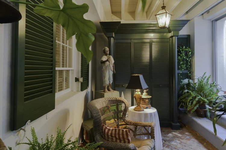 Side view of porch decorated with pale wicker furniture, dark green shutter closet, and many ferns and potted plants