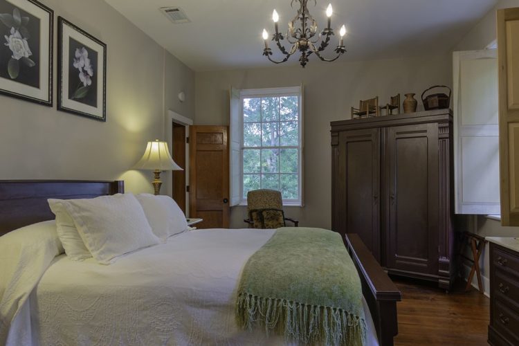 Side view of a bedroom decorated with a large bed with a white bedspread and green throw blanket, dark wooden dressers, and two bedstands with lamps