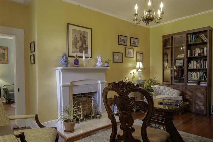 View of sitting area decorated with lemon yellow walls, tan and beige chairs, a white and brick fireplace, wood flooring, and a rustic bookshelf