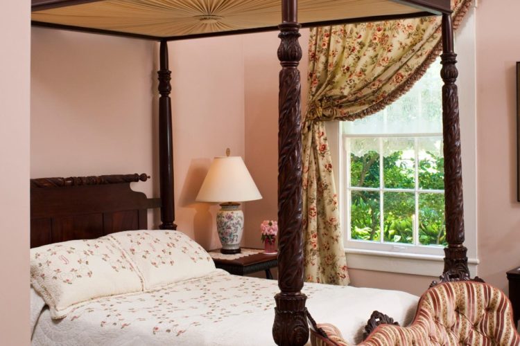 Bedroom decorated with a dark wooden poster bed covered in a white flowery bedspread, a window with flowery curtains, and pale pink walls