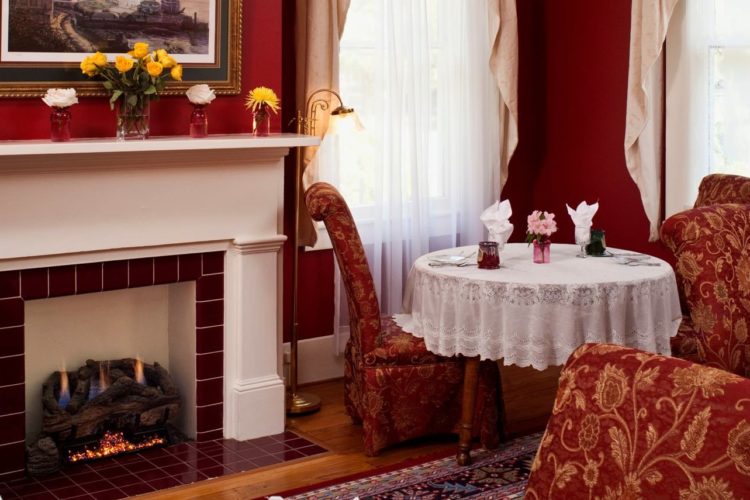 Sitting area decorated with crimson red walls, red and gold patterned chairs, a white and red tiled fireplace, and white lace tablecloth over round table