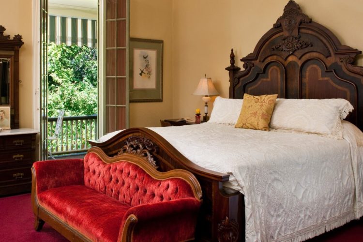 Vintage bedroom decorated with elegant dark wood bedframe and white comforter, a red victorian sofa, wooden dresser and bedstands, red carpet, and cream walls