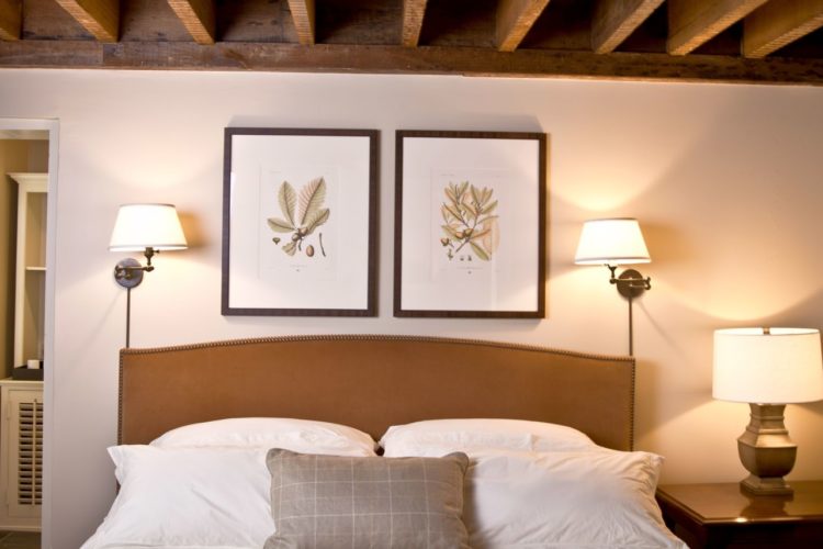Low-ceiling bedroom with queen-sized bed and white pillows, a small wood desk in corner with a lamp, two bed lamps, and two picture frames