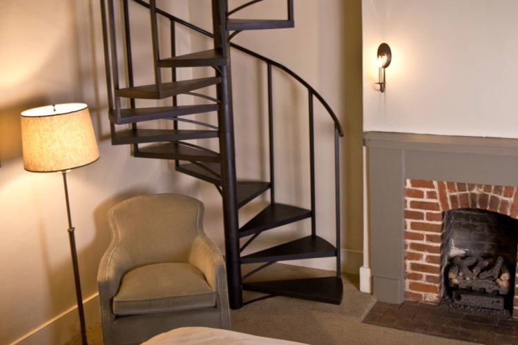 Sitting area decorated with cream walls, a warm gray chair and brick fireplace, and dark brown spiral staircase in the back corner