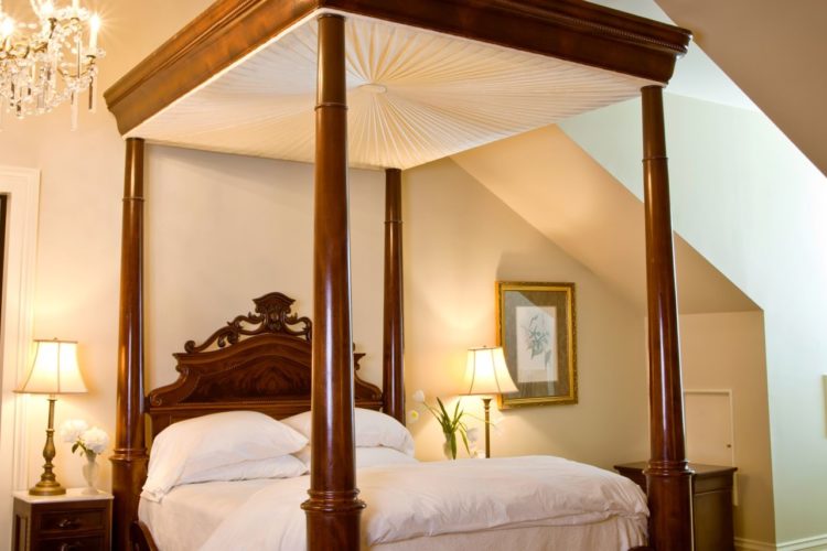 Cream-colored bedroom with tall wooden poster bed and white linens, two bedstands with flowers and lamps, and an alcove with a window