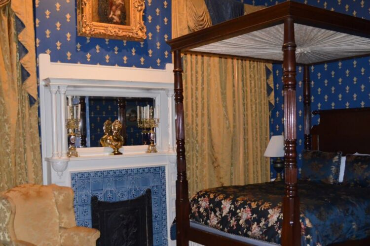 Intricate bedroom with royal blue and gold accented wallpaper, blue-tiled fireplace, silk navy blue and pink-flowered bedspread on poster bed, and golden curtains and chairs