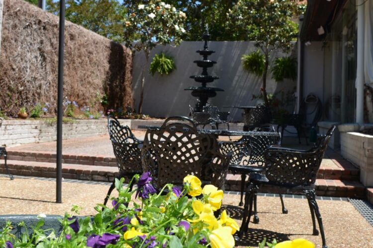 Outdoor seating area with black porch furniture, purple and yellow flowers, tall black water fountain in background and walled in with trees behind