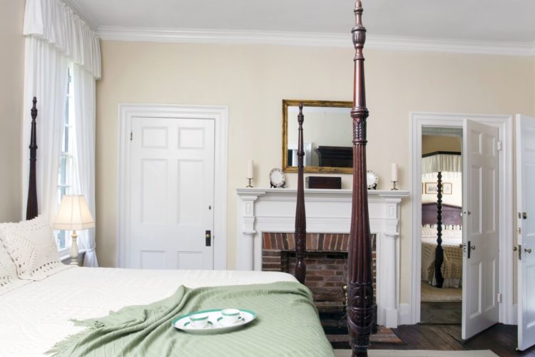 Cottage style bedroom decorated with eggshell walls, dark wood flooring, a king-sized bed with white linens and green throw blanket, a brick fireplace, and three white doors
