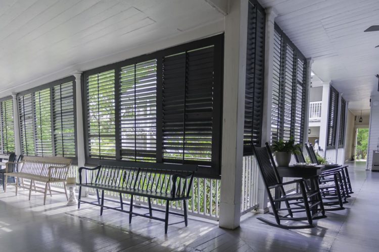 Large covered porch area painted bright white with black shutters for weather protection, multiple benches and rocking chairs overlooking greenery