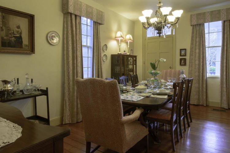 Dining room with pale yellow walls, wood flooring, dark wood dining table with eight chairs, two large windows and one door, and dish cabinet against wall