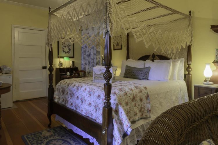 Bedroom scene depicting pale yellow walls, wood flooring, a large poster bed with white quilted linens, and dining table to the left