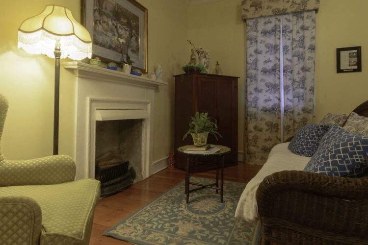 Sitting area with pale yellow walls, wood flooring with baby blue accented rug, white fireplace, wicker loveseat, and yellow reading chair