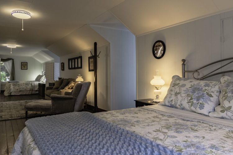 Large white bedroom consisting of two beds with flower print quilts, a sitting area between them with gray chairs and a flatscreen television