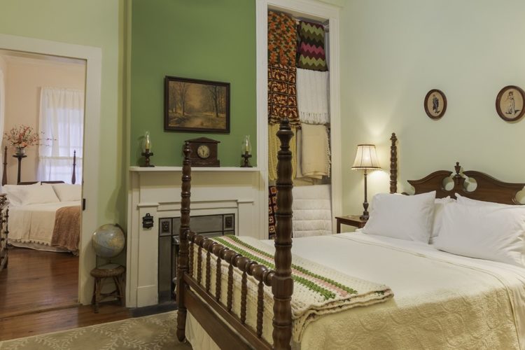 Light green styled bedroom with queen-sized bed and cream comforter, white fireplace, two bedstands and lamps with door leading to separate bedroom