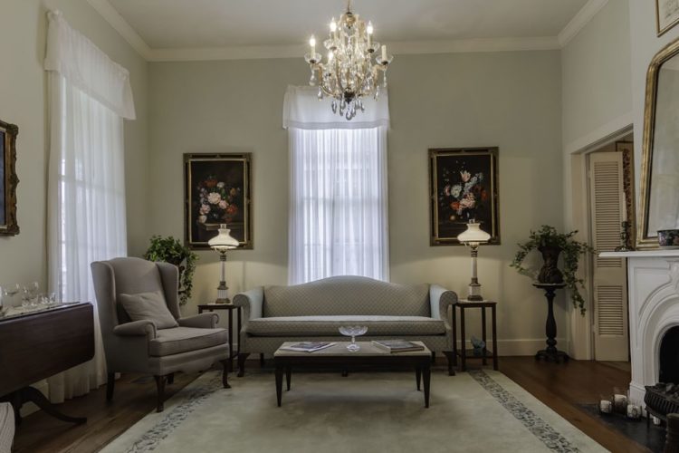 Neat living room area with cream walls, dark wood flooring and cream carpet, light gray furniture, flower paintings, and white fireplace with small coffee table