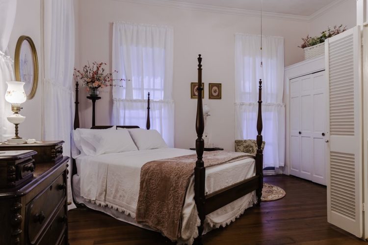White bedroom decorated with a dark wooden dresser with two antique lamps, dark wood flooring, a bed with a white bedspread and cocoa throw blanket, and white cabinetry