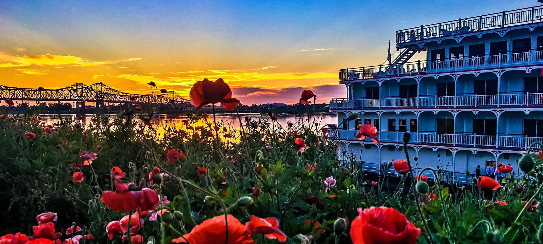 Blue, orange, and pink sky with sun setting behind a bridge near a river with a white riverboat and red flowers near the edge of the river